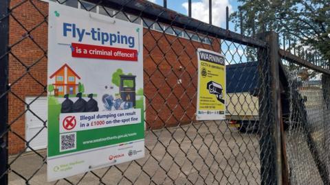A close up shot of a wire fence, with a white poster strapped on. It says "Fly-tipping is a criminal offence. Illegal dumping can result in a £1000 on-the-spot fine. www.westnorthants.gov.uk/waste". There is another yellow poster warning people that CCTV is in operation.