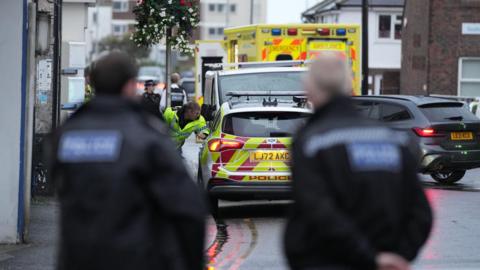 A police officer looking into the passenger window of a police car parked on the left hand side of the road, an ambulance is parked in front of the police car. Two police officers are stood closer and with their backs to the camera. They are blurred in the image.