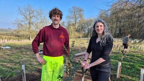 Two people stand together in a field of young trees being planted.