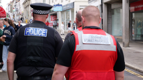 Three men from behind, one wearing a police uniform and two wearing red Bodmin street marshal vests patrolling a town street with people in front of them.