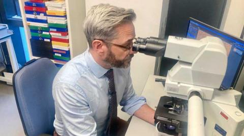 Children’s Brain Tumour Centre of Excellence co-director, Professor Richard Gilbertson, has greying hair and a beard and wears brown framed glasses. He is wearing a light blue shirt with a brown spotted tie and is looking through a microscope.