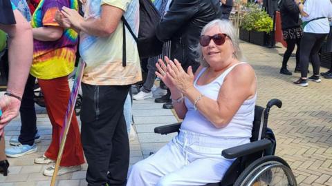 Glynis Bell is pictured in her wheelchair during a Pride event. She smiles at the camera while clapping her hands. She wears sunglasses with a white vest top and loose white trousers. She has short grey hair.