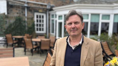 Deputy Andy Sloan has grey and black hair and is smiling at the camera. He's wearing a brown jacket on top of a blue polo shirt and a white polo shirt. He's standing amongst a load of tables at an outdoor restaurant.