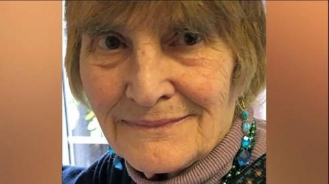 Close-up image of Patricia Holland wearing a polo-neck jumper with a beaded necklace. She is sat in front of a window.
