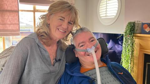 The image shows a man and a woman positioned closely together indoors. The man is seated in a supportive chair, with a visible ventilation tube running from the lower part of the frame towards his chest. He is wearing a padded grey top with a blue outer layer. The woman is leaning in beside him and is wearing a textured long‑sleeved shirt with a microphone clipped to the front. Behind the two people is a round window with shutter-style blinds, and a fireplace with a wooden mantel.