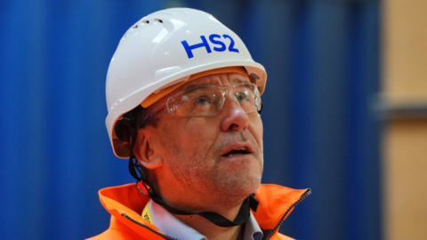A man wearing a white helmet marked HS2 and orange hi-viz overalls