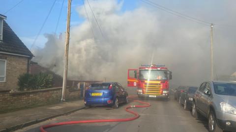 A fire engine parked on a smoke-filled residential street. Thick grey smoke rises behind the houses and a large red hose runs from the fire engine along the road past parked cars.