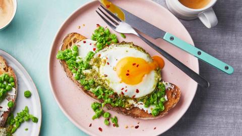 Pesto and a fried eggs on a slice of sourdough toast on a pink plate with a knife and fork.