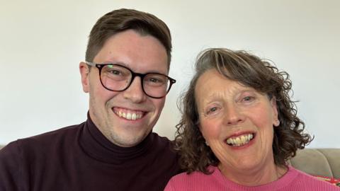 A younger man and an older woman sitting together on a beige sofa smiling at the camera. The man on the left wears black-rimmed glasses. The woman has frizzy brown hair and is wearing lipstick.