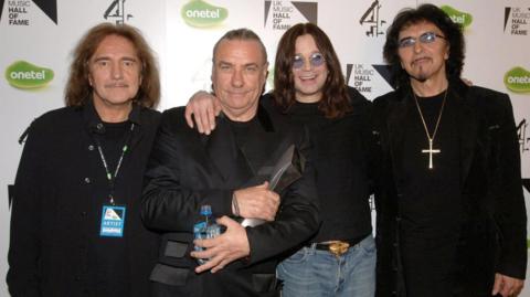 Four men wearing dark clothing standing in a line with their backs against an advertising board. From left to right are Black Sabbath band members Geezer Butler, Bill Ward, Ozzy Osbourne and Tony Iommi.