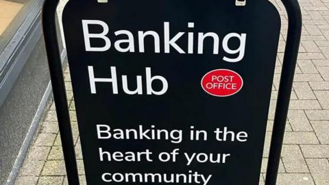 A black sign on a street with white writing that says "banking hub, banking in the heart of your community"