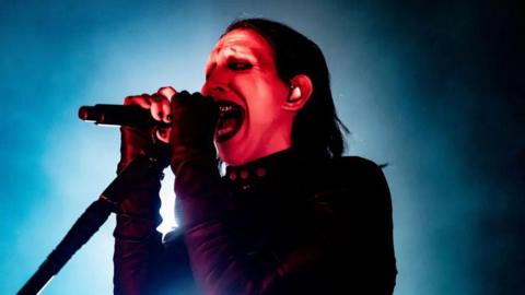 Close up image of Marilyn Manson on stage singing in full make up holding a microphone