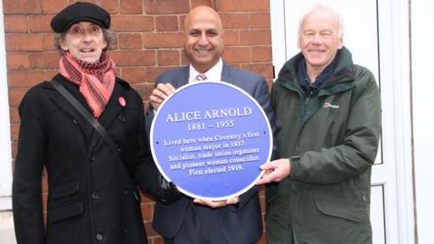 Three men standing holding a large blue circular plaque, with the name Alice Arnold written at the top.