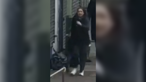 A blurry CCTV image of a woman in black trousers and a black jacket walking down the street. She is wearing white trainers. 