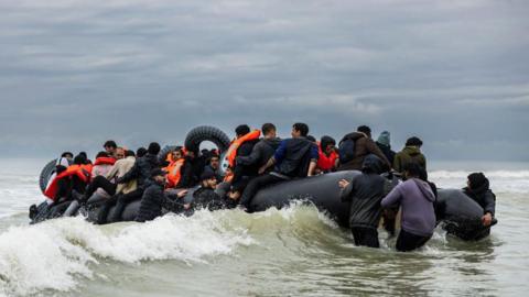 A raft of people - some in orange life jackets - struggles in the waves. Some of the men are in the water, pushing the vessel.