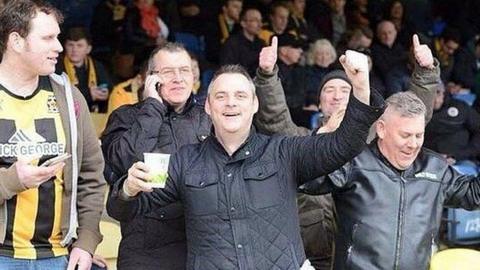 Simon Dobbin in the stand at a football stadium and holding a cup of tea. He has a fist in the air and is celebrating. He has short greying hair and is wearing a black coat in front of other football supporters.