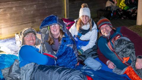 Four people inside sleeping bags. They are looking at the camera and smiling. There is a wood wall behind them. There are wearing hats and coats. 