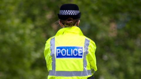 The back of a woman police officer, who has a hi-vis police coat on with "police" on the back of it and is wearing a woman's police hat as she faces large trees or bushes 