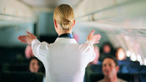 An air stewardess facing away from the camera shows passengers on a jet the emergency exist during a safety briefing. She is wearing a white shirt and her blonde hair is in a tight bun