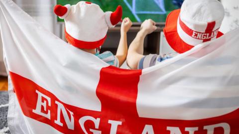 Two young England fans drapped in a ST George's cross cheer as they watch the Three Lions play on tv.