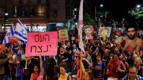 An overhead shot of a crowd of protesters taken at night-time. Some hold up posters with images of hostages, others hold flags and others hold up signs. The crowd appears to be progressing forwards.