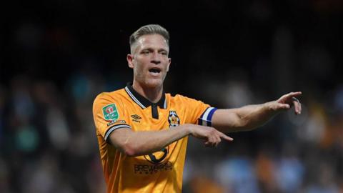 Morrison pointing with both arms as he hands out instructions, wearing gold and black home kit of Cambridge United