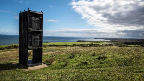 The Pit Cage Monument stands on a grassy field with the coast visible in the distance. It is a sunny day with a clear sky over the sea and clouds towards land.