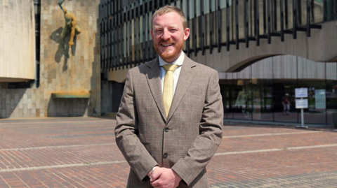Colin Ferguson has short ginger hair and a beard. He stands outside the civic centre in Newcastle smiling.