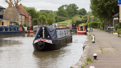 About five narrowboats on a canal, with a path on the right. There are buildings to the left and right, and trees behind the boats. A few people can be seen on the boats. 