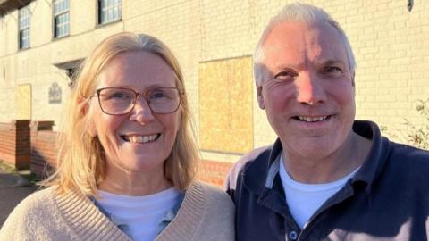 Debbie, on the left, and Mike on the right. She is wearing a beige pullover, and white teeshirt beneath. She wears glasses, and has long shoulder-length blonde hair. Mike has greying hair, slightly receding, and is wearing a blue polo shirt and white teeshirt beneath. Both are smiling, standing in front of the fire-damaged Gables Farm, which they have purchased.