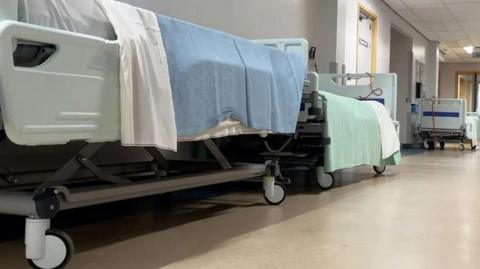 Empty hospital beds line the corridor of a hospital ward. They have blue and green blankets draped across them.