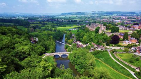 An aerial drone shot looking over Ludlow. You can see the River Severn with an arched bridge going over it. To its right is Ludlow Castle, with green ;and and houses, and the town in the distance. In the far background are more houses and rolling green hills and fields