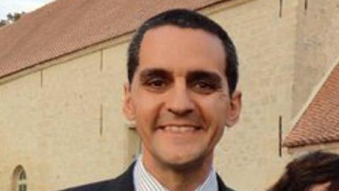 Richard Osman, who has short dark hair and is wearing a striped shirt and suit jacket and is smiling at the camera. A beige coloured building is behind him.