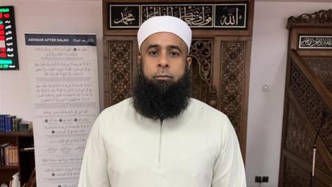 A man with a long black beard wearing a white long sleeve top. He is wearing a white linen hat on his head. He is stood in front of a wall hanging with Arabic test on the top of it.