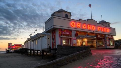 Weston-super-Mare pier is pictured at sundown. At the entrance the words 'Grand Pier' stand out in almost a neon orange/red colour. That light is reflected on the cobbles in front of the entrance. The beach and sea can also be seen and there are dark clouds passing overhead. 