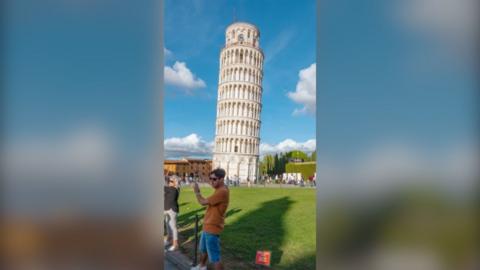 A man wearing an orange t shirt and blue shorts is stood in front of the Leaning Tower of Pisa, with his hands raised up pretending to hold it from falling.