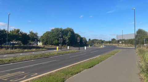 An empty road with trees along the left hand side and a set of traffic lights in the middle of the shot