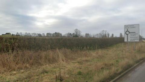 The side of a road with a hedgerow and trees and a couple of houses in the distance. A road sign for a roundabout directing drivers to the town centre, Thirsk, or Darlington, Richmond or industrial estates.