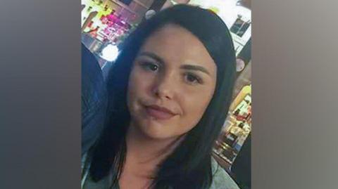 Alice Webb is smiling at the camera, she has dark hair and a piercing in her lower lip. She stands in front of an illuminated bar.