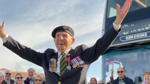 An old man wearing a military uniform and medals stands smiling with his arms outstretched in front of a bus. People are standing and smiling in the background. 