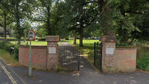 A corner entrance to the park in North Walsham, featuring two sections of curved red brick wall either side of a black wrought iron gate. Beyond the gate, a concrete path goes off into the distance, lined by tall trees with lawns either side.
