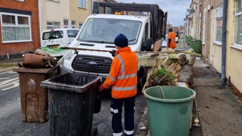 A council worker in a high vis jacket and work trousers with stripes is loading up bins onto a council truck.  The vehicle is parked on a street of terraced houses.  One resident is watching from his doorway.