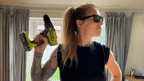 A woman in sunglasses holds up a power drill.