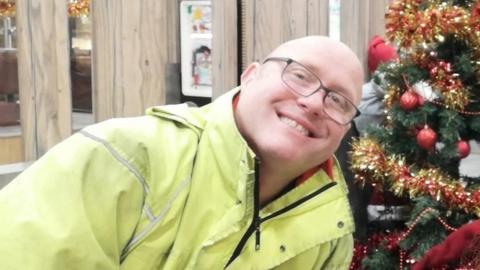 Andrew Farnsworth in yellow Royal Mail jacket. He has a shaved head and is wearing dark glasses.