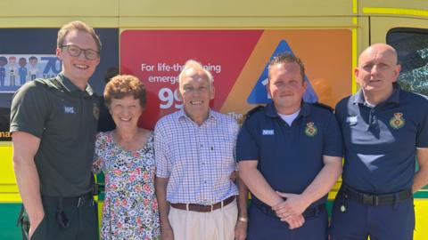 Five people standing in front of an ambulance. Including paramedic on the left wearing green uniform and two first responders on the right wearing dark blue uniform.