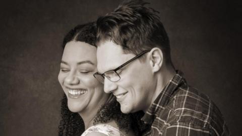A black and white image of David Clark, wearing glasses and a plaid shirt, embracing his wife.