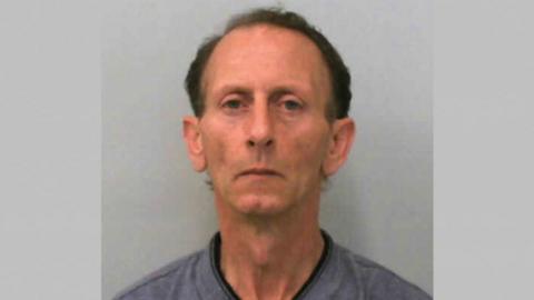 A police mugshot of Ricky Morgan, wearing a dark grey shirt and standing against a grey background. He has dark brown hair and is clean shaven, looking at the camera with a blank expression.