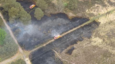An aerial view of the fire being put out by two engines which are parked on the land and on a path. There is water spraying from the side of one engine and the plants on the ground are black with smoke rising up
