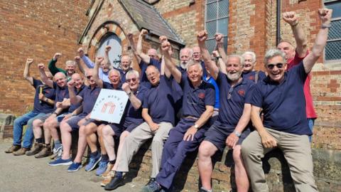 A group of older men gathered outside a brick building with arched windows, possibly a church or community centre. They wear matching navy blue shirts with a logo and pose cheerfully, some raising their fists in celebration. One man at the front holds a sign reading “Ripon Men’s Shed” with a red logo.