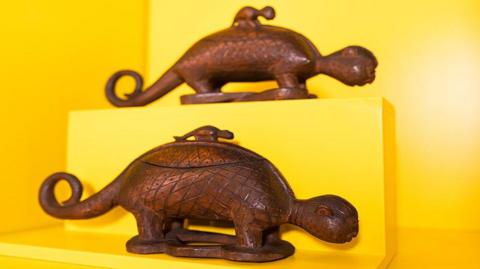 Two carved wooden animals on show on a yellow exhibition display shelf.